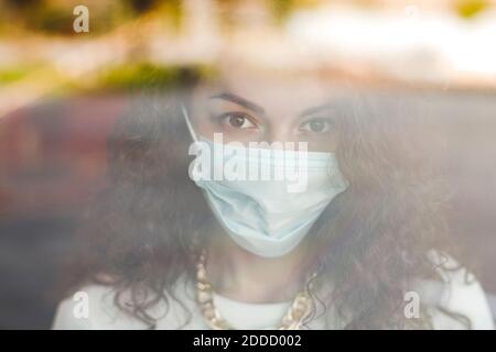 Belle jeune femme portant un masque protecteur vu à travers le verre Fenêtre pendant la pandémie de COVID-19 Banque D'Images