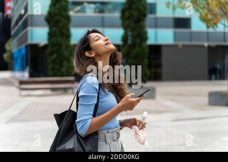 Belle femme avec masque de protection et assainisseur tenant intelligemment Téléphone en ville pendant COVID-19 Banque D'Images