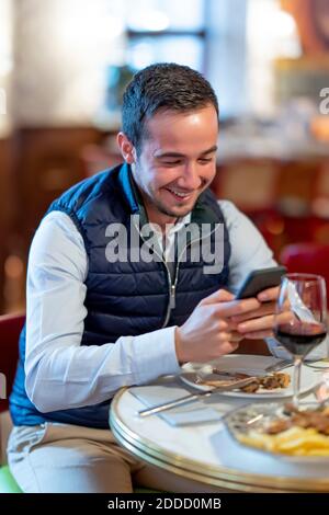 Un jeune homme heureux avec messagerie texte sur un smartphone à la table de salle à manger du restaurant Banque D'Images