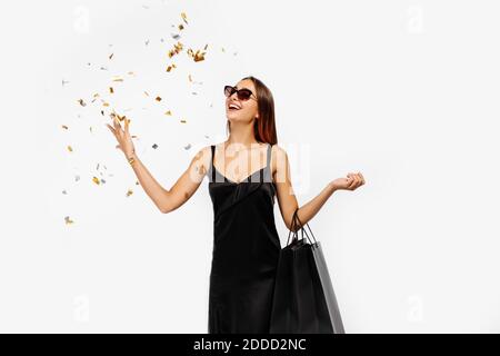 Jeune femme excitée, en robe noire, avec des sacs de shopping, célébrant et jetant des confetti sur fond blanc, concept, shopping, Black Friday Banque D'Images