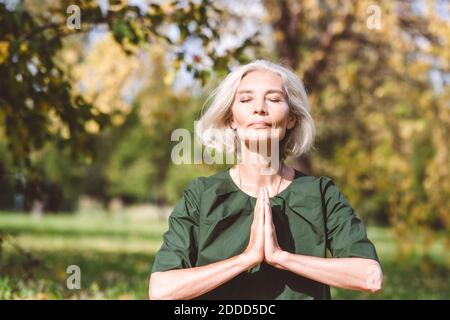 Femme mûre assise avec les mains jointes tout en faisant le yoga à stationnement Banque D'Images
