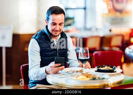 Un jeune homme souriant vous envoyer des SMS sur un smartphone en étant assis dans le restaurant Banque D'Images