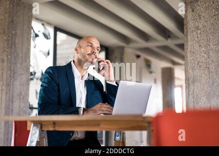 Homme d'affaires vêtu d'un costume qui parle sur un téléphone portable tout en travaillant ordinateur portable au bureau Banque D'Images
