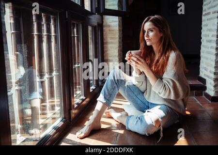 Une jeune femme boit du café tout en étant assise sur le sol à la maison Banque D'Images