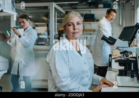 Femme scientifique assise au microscope avec un collègue travaillant en arrière-plan au laboratoire Banque D'Images