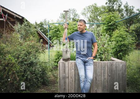 Beau homme tenant la bêche en regardant loin tout en s'appuyant sur le bois lit surélevé au potager Banque D'Images