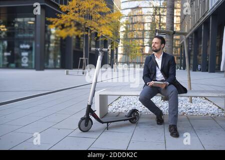 Homme d'affaires avec tablette numérique assis par e-scooter sur le banc ville Banque D'Images