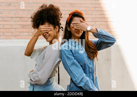 Des amies féminines couvrant les yeux tout en se tenant dos à dos contre mur Banque D'Images