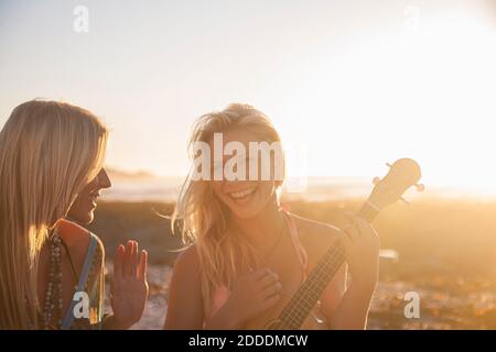 Jeune femme gaie jouant ukulele pendant que l'amie chantait à plage au coucher du soleil Banque D'Images