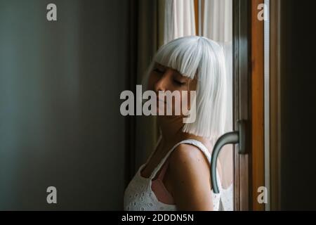 Jeune femme avec les yeux fermés penchée sur la fenêtre à la maison Banque D'Images