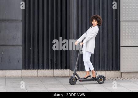 Femme d'affaires excitée à la conduite d'un scooter électrique contre le mur Banque D'Images