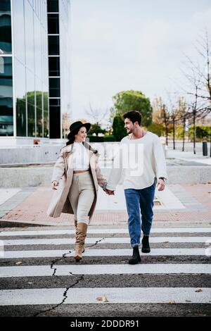 Belle femme souriante tenant les mains avec un partenaire mâle pendant la traversée route en ville Banque D'Images