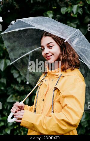 Fille souriante en imperméable tenant l'appareil photo tout en se tenant contre la feuille mur Banque D'Images