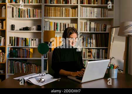 Femme d'affaires souriante travaillant sur un ordinateur portable, assise contre une étagère à la maison Banque D'Images