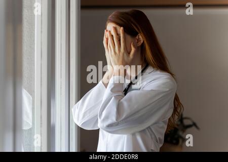 Jeune femme médecin émotionnellement stressée avec la tête dans les mains debout par fenêtre Banque D'Images