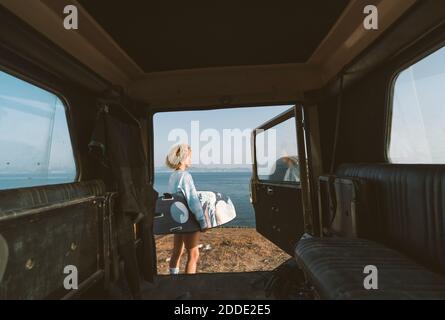 Jeune femme tenant une planche de surf tout en se tenant en voiture contre la mer à la plage Banque D'Images