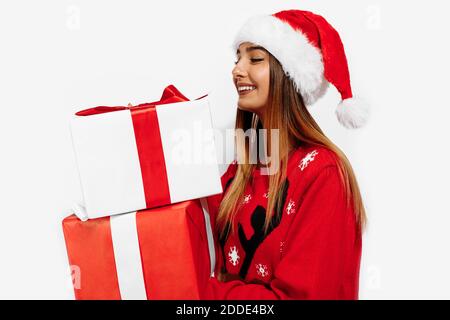 Jeune femme excitée en chandail rouge et chapeau de père noël tenant de nombreux cadeaux sur fond blanc, noël, concept de vacances Banque D'Images