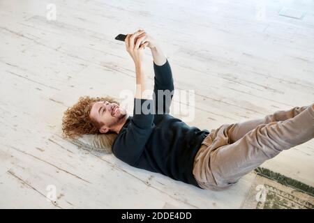 Homme souriant prenant le selfie à travers un smartphone couché sol à la maison Banque D'Images