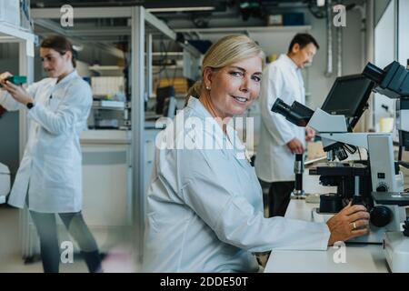 Scientifique souriant assis au microscope avec des collègues travaillant en arrière-plan au laboratoire Banque D'Images