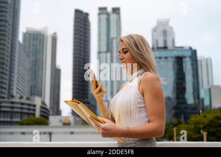 Belle femme d'affaires blonde utilisant un téléphone mobile tout en tenant le dossier à centre-ville Banque D'Images