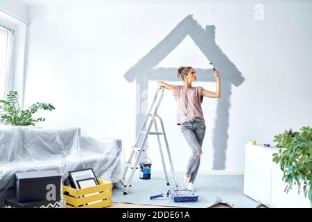 Jeune femme avec rouleau de peinture faisant pout tout en s'appuyant sur échelle à la maison Banque D'Images
