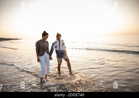 Un jeune couple heureux tient les mains tout en marchant sur la rive à plage au coucher du soleil Banque D'Images