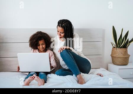 Fille utilisant un ordinateur portable tandis que la mère s'assoit à côté d'elle sur le lit dans la chambre à la maison Banque D'Images