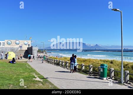 Table Mountain, Devils Peak et Lions Head vus depuis la plage de Bloubergstrand, de l'autre côté de Table Bay. Les gens marchent le long de la plage et s'assoient sur l'herbe. Banque D'Images
