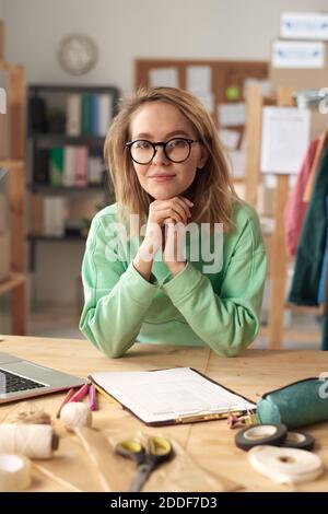 Portrait de jeune tailleur dans les lunettes regardant l'appareil-photo pendant assis sur son lieu de travail Banque D'Images