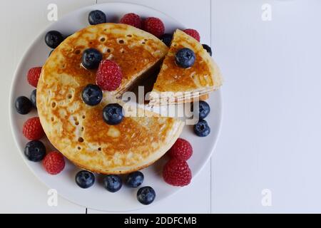 Assiette de crêpes coupée en triangle avec sirop d'érable, bleuets et framboises sur table en bois blanc et vue de dessus. Banque D'Images