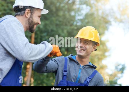 Deux jeunes ingénieurs masculins positifs en casques durs souriant l'un à l'autre, donnant le coup de poing tout en travaillant sur le chantier de construction de chalet à l'extérieur. Construction, travail d'équipe, concept de partenariat Banque D'Images