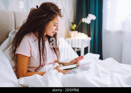 La jeune femme aux cheveux afro fait ses achats en ligne en utilisant son smartphone assis à la maison dans le lit payant avec carte bancaire Banque D'Images