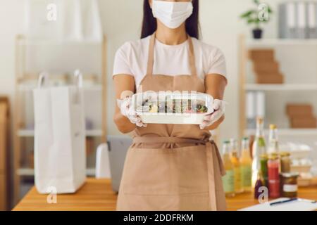 Un employé de café à emporter dans un masque médical tenant un contenant déjeuner prêt pour la livraison Banque D'Images