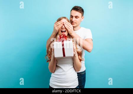 Un beau homme ferme les yeux de sa femme, lui faisant une surprise, et lui donne un cadeau sur fond bleu. Saint Valentin Banque D'Images