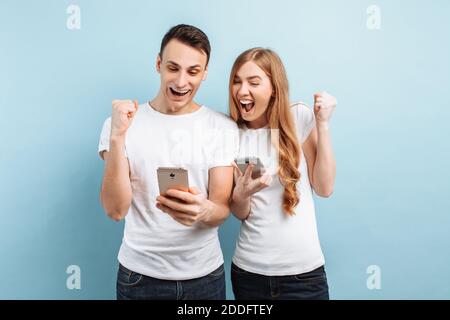 Photo de personnes enthousiastes, hommes et femmes, en T-shirts blancs, montrant avec joie un geste de victoire et de succès, tenant les smartphones dans leurs mains, encore une fois Banque D'Images