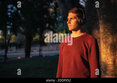 Un garçon de race blanche blonde qui écoute de la musique avec un casque sans fil au coucher du soleil, portant un chandail rouge. Concept de technologie. Musique dans la rue. Banque D'Images