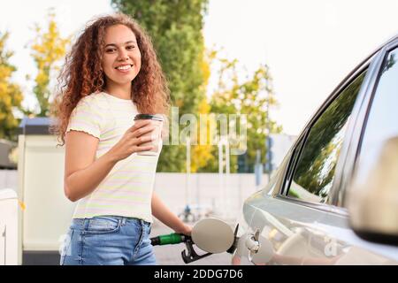 Femme afro-américaine qui boit du café tout en remplissant le réservoir de voiture à station-service Banque D'Images