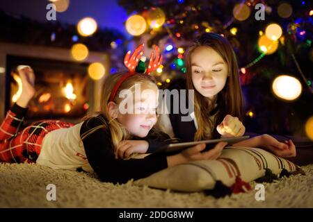 Deux jolies jeunes sœurs à l'aide d'un tablet pc à la maison par une cheminée dans le salon chaleureux et confortable, la veille de Noël. Soirée d'hiver à la maison avec la famille et Banque D'Images