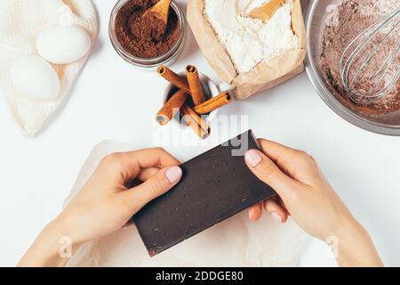 Les femmes tiennent une barre de chocolat noir à côté des ingrédients pour faire cuire du brownie maison avec de la cannelle sur une table blanche, vue du dessus. Banque D'Images