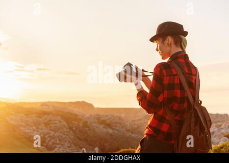 Vue latérale d'une femme voyageur androgyne avec appareil photo professionnel debout sur une colline dans une zone montagneuse et regardant à travers pris photos Banque D'Images