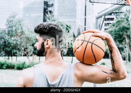 Vue arrière d'un jeune homme à barbe plein de confiance avec tatouage bras tenant le ballon de basket-ball et regardant loin tout en restant debout aire de jeux Banque D'Images