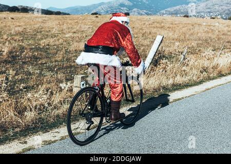 Vue latérale du père Noël en costume rouge moderne faites du vélo sur une route vide dans les hauts plateaux par beau temps Banque D'Images