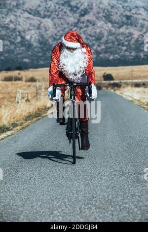 Le Père Noël en costume rouge à vélo moderne le long de vide route dans les hauts plateaux par jour ensoleillé Banque D'Images