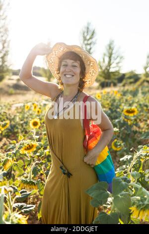 Vue latérale d'une femme sereine avec sac arc-en-ciel debout champ de tournesol en plein essor et beau temps Banque D'Images