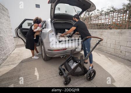 Vue arrière de l'homme ethnique qui porte des bagages et poussette de bébé Dans le tronc et une mère afro-américaine avec un tout-petit qui entre voiture pendant la préparation du voyage Banque D'Images