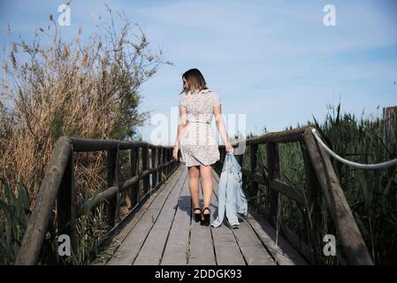 Vue intégrale sur le dos de la jeune femme en robe décontractée avec une veste à la main se promenant sur une passerelle en bois parmi les hauts herbe sèche par beau temps Banque D'Images