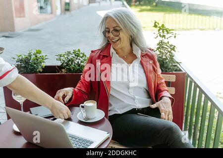 Serveur de récolte servant du café pour une femme entrepreneure mûre et positive assise à la table dans un café extérieur Banque D'Images