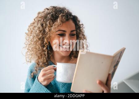 Une femme joyeuse dans un confortable chandail domestique penchée sur les escaliers à maison tout en appréciant l'histoire intéressante et en buvant un café chaud à week-end Banque D'Images