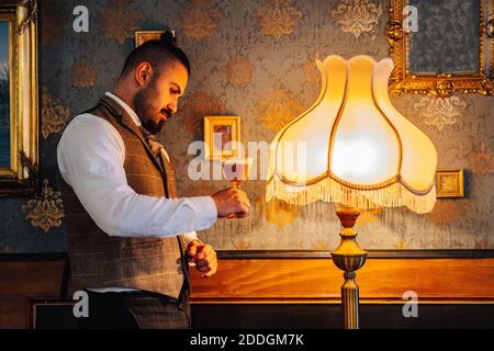 Vue latérale d'un homme sérieux et réfléchi portant un verre de Café irlandais près d'une lampe à l'ancienne dans une chambre classique Banque D'Images