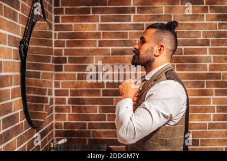 Vue latérale d'un homme masculin à barbe en tenue habillée régler l'attache et regarder la réflexion dans le miroir Banque D'Images